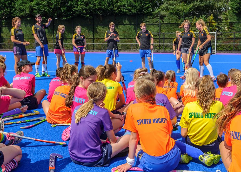 A large group of young players sitting on a blue pitch listening to a coach